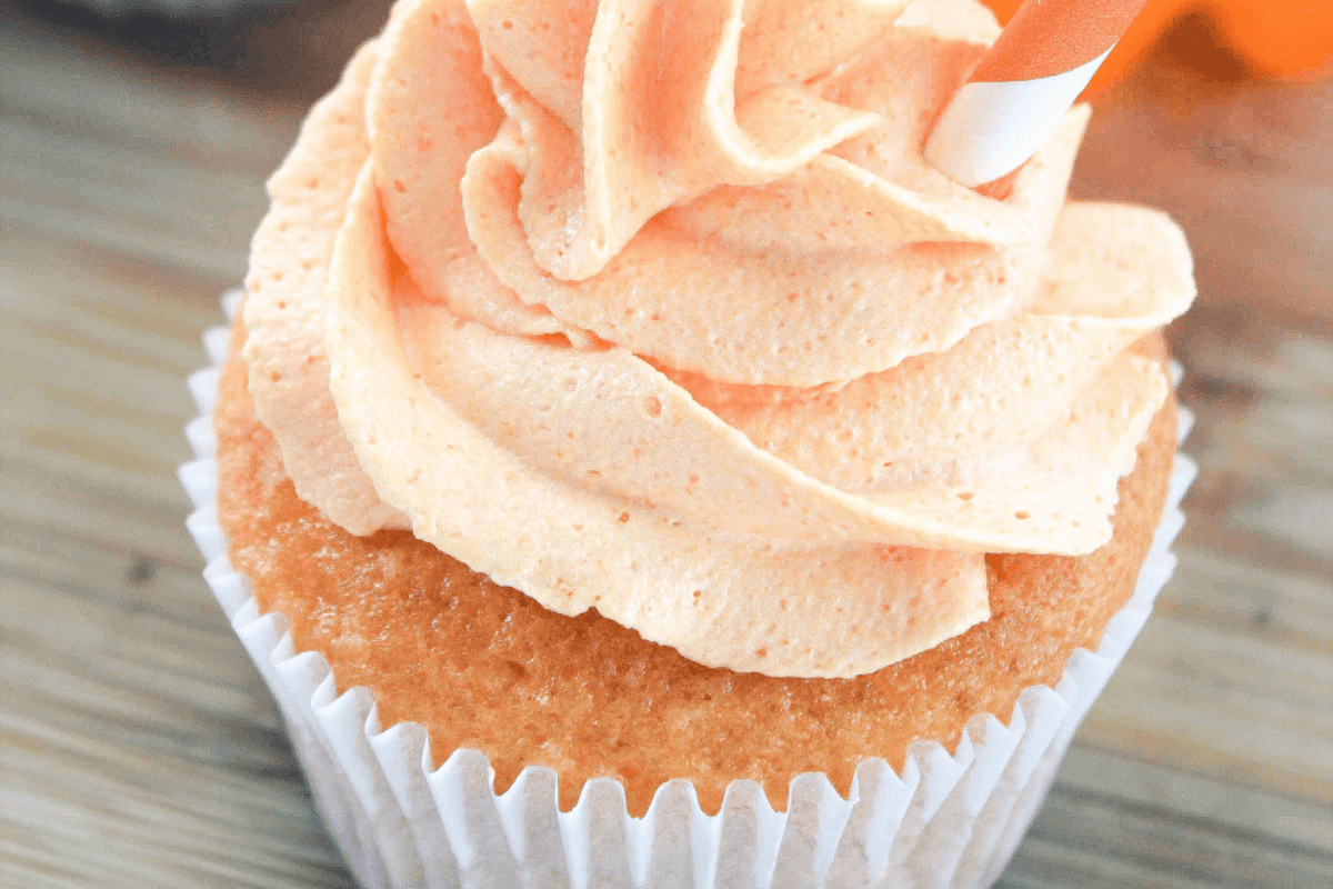 A close-up of a cupcake with light orange frosting swirled on top, set in a pale paper cupcake liner. The frosting is smooth and peaks attractively. The background features a wooden surface, slightly blurred.