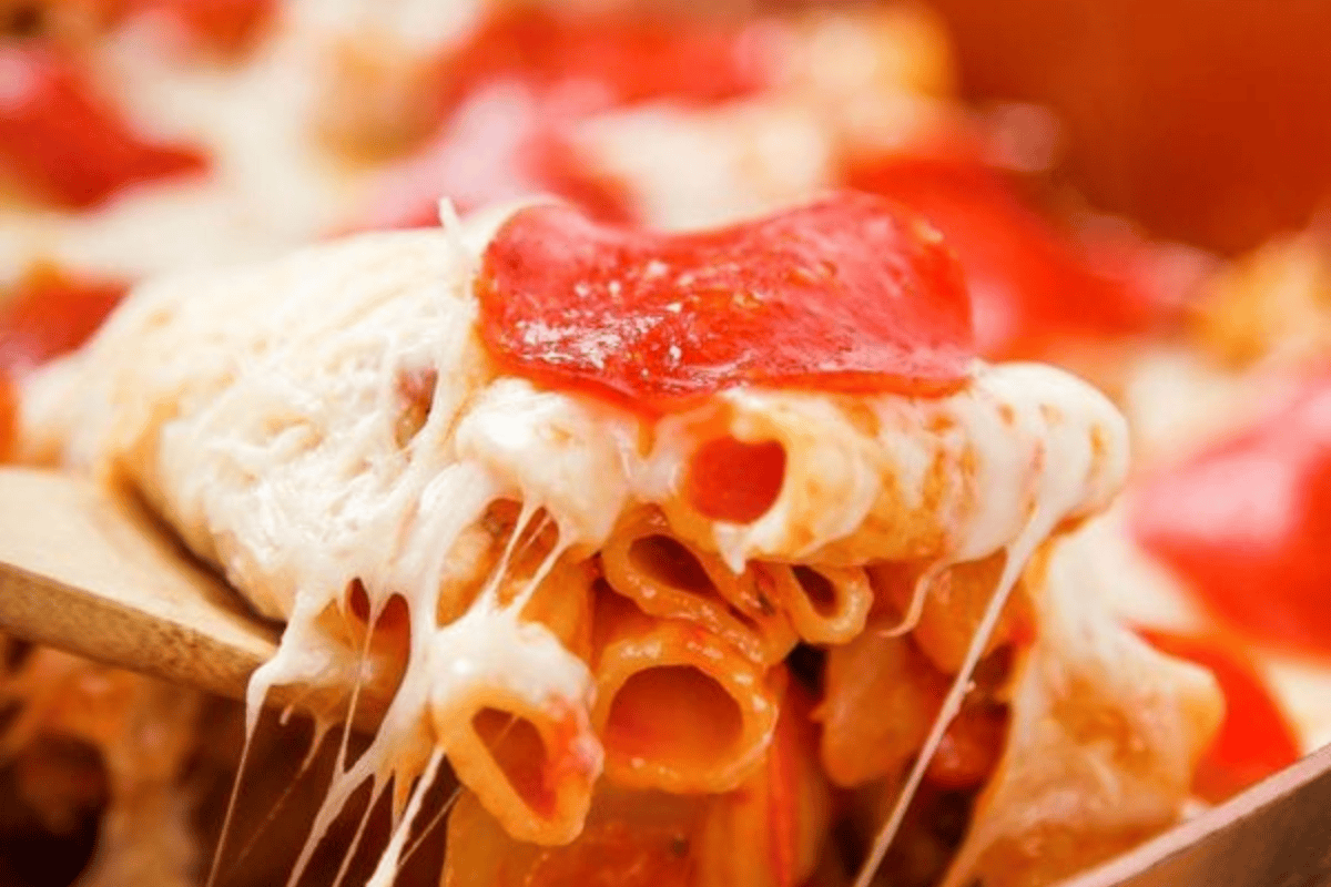 Close-up of a cheesy baked pasta dish being scooped with a wooden spoon. The pasta is topped with melted mozzarella cheese and pepperoni slices, with strings of gooey cheese stretching as the pasta is lifted. The colors are warm and inviting.