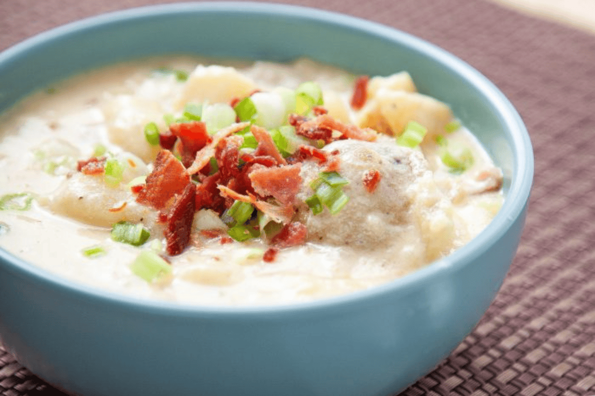 A blue bowl filled with creamy potato soup topped with crispy bacon pieces and chopped green onions. The soup appears thick and hearty, featuring chunks of potatoes and meat. The bowl is placed on a woven placemat.