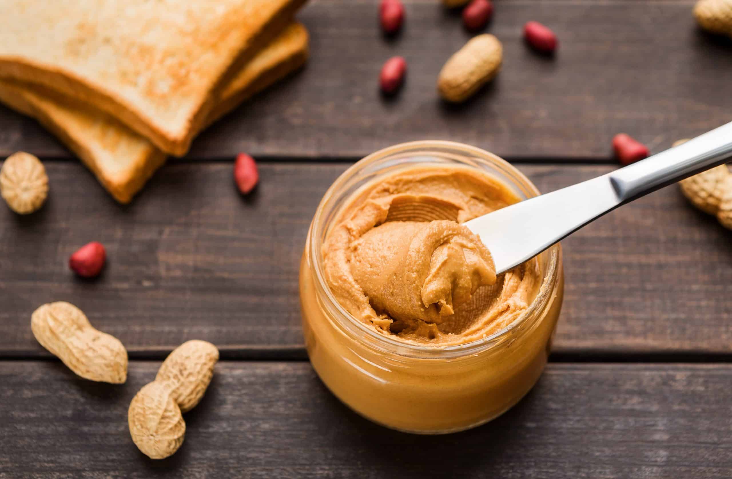 Peanut butter in jar, toasts and scattered peanuts on wooden background. Vegetarian butter concept