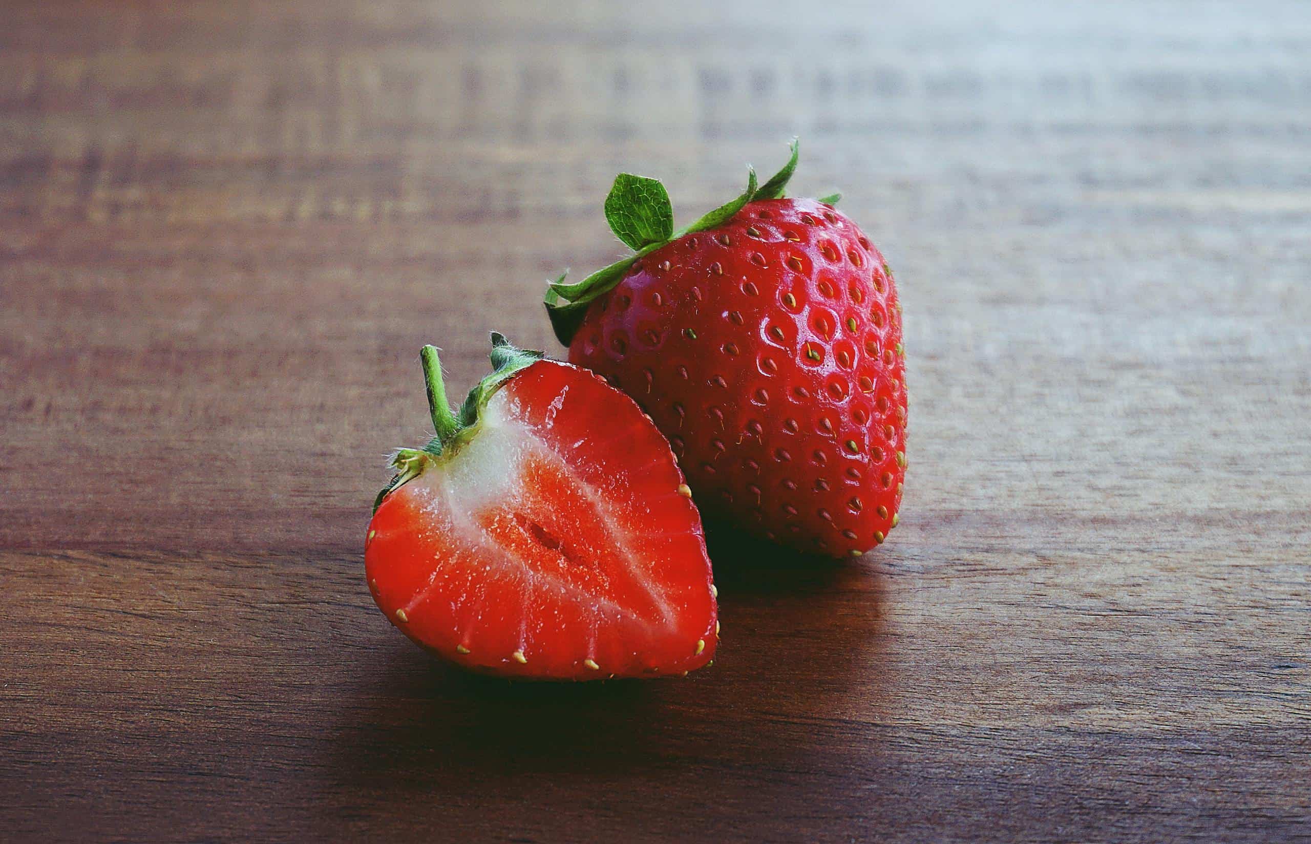 Two fresh strawberries, one whole and one sliced, on a wooden surface.