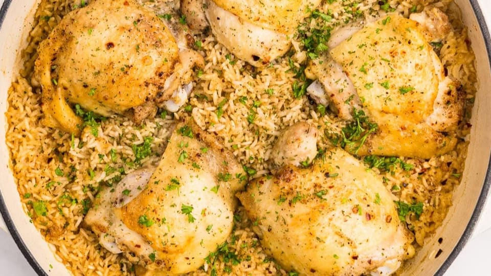A white pot containing browned chicken thighs sitting in a pot full of broth and rice before baking.