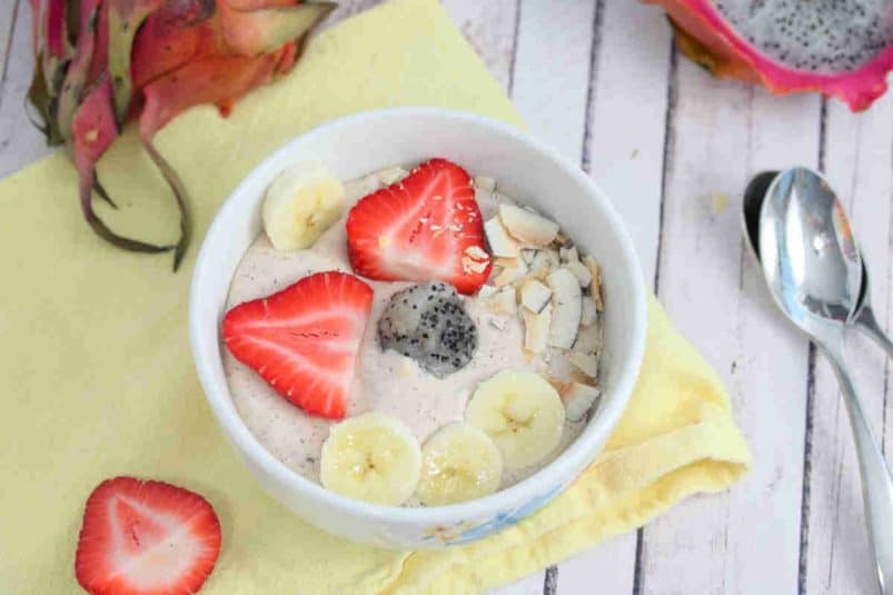 White bowl with a dragon fruit smoothie bowl in it topped with bananas, strawberries, and coconut on a yellow linen with berries, dragon fruit, and spoons around it.