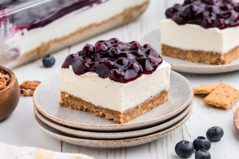 No bake cheesecake bar on a plate with blueberry pie filling on top. Another slice in the background.