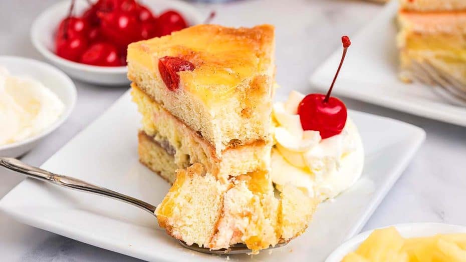 A slice of pineapple upside-down cake on a plate, garnished with whipped cream and a cherry, with a fork resting beside it.