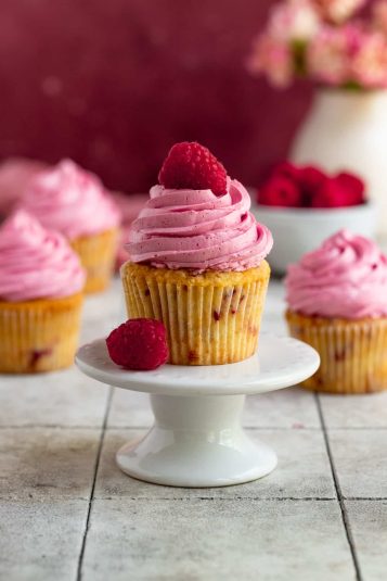 A single raspberry cupcake with raspberry buttercream on a mini cake stand. The cupcake is topped with a fresh raspberry. There are three cupcakes in the background and a small bowl of fresh raspberries.