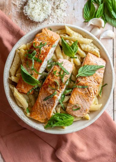 Creamy penne pasta topped with pan-seared salmon filets and garnished with fresh basil. The bowl is on top of a pink linen napkin.
