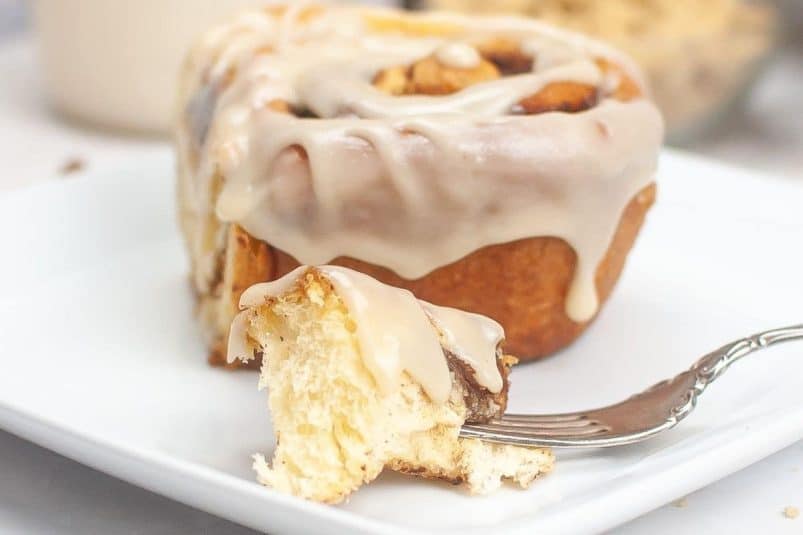 A close-up of a sourdough cinnamon roll with white icing on a white plate. A bite has been taken out, and a piece is on a fork in front.