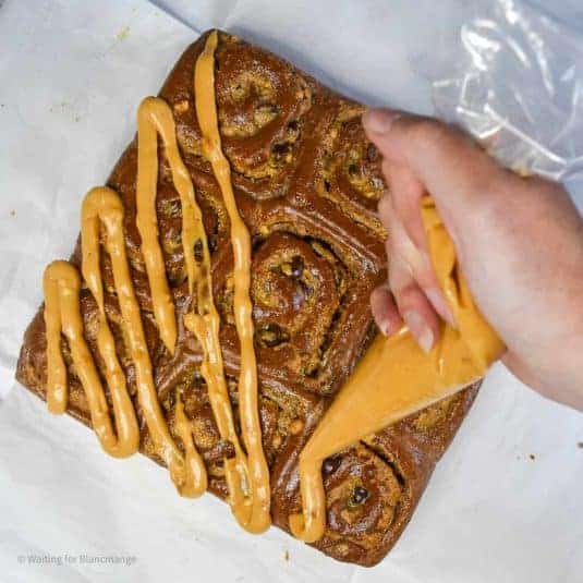 An overhead image of dark chocolate rolls with peanut butter and chocolate chip filling. The rolls have been removed from their pan and are sat on a white countertop on top of parchment paper. There is a hand holding a piping bag over top of them, piping a zig zag of thick peanut frosting.