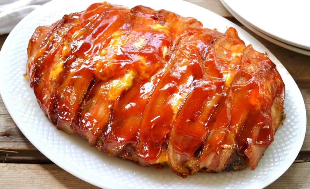 Meatloaf wrapped in bacon and glazed with ketchup on a white platter.