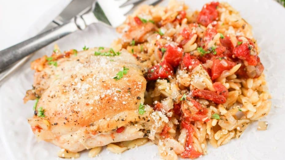 A plate with baked chicken thigh and orzo pasta mixed with tomatoes, garnished with herbs, with a fork and knife beside.
