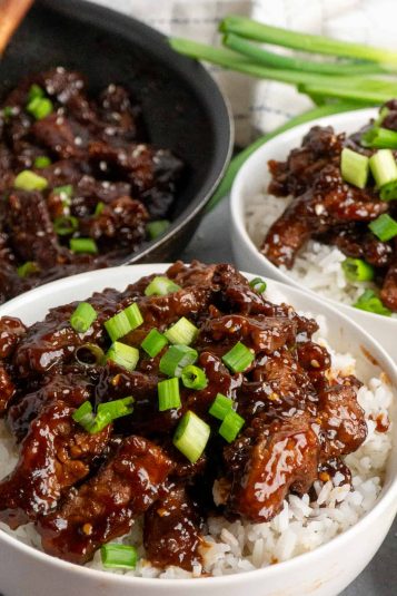 Beef teriyaki over a bowl of rice with a pan of more beef in the back and some stalks of green onions.