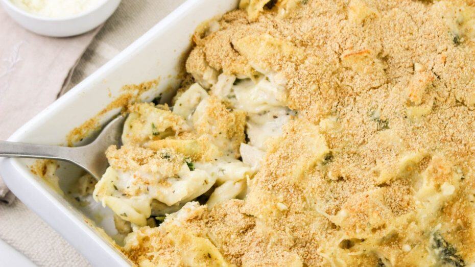 A baking dish filled with creamy pasta topped with a golden breadcrumb crust. A spoon is serving a portion.