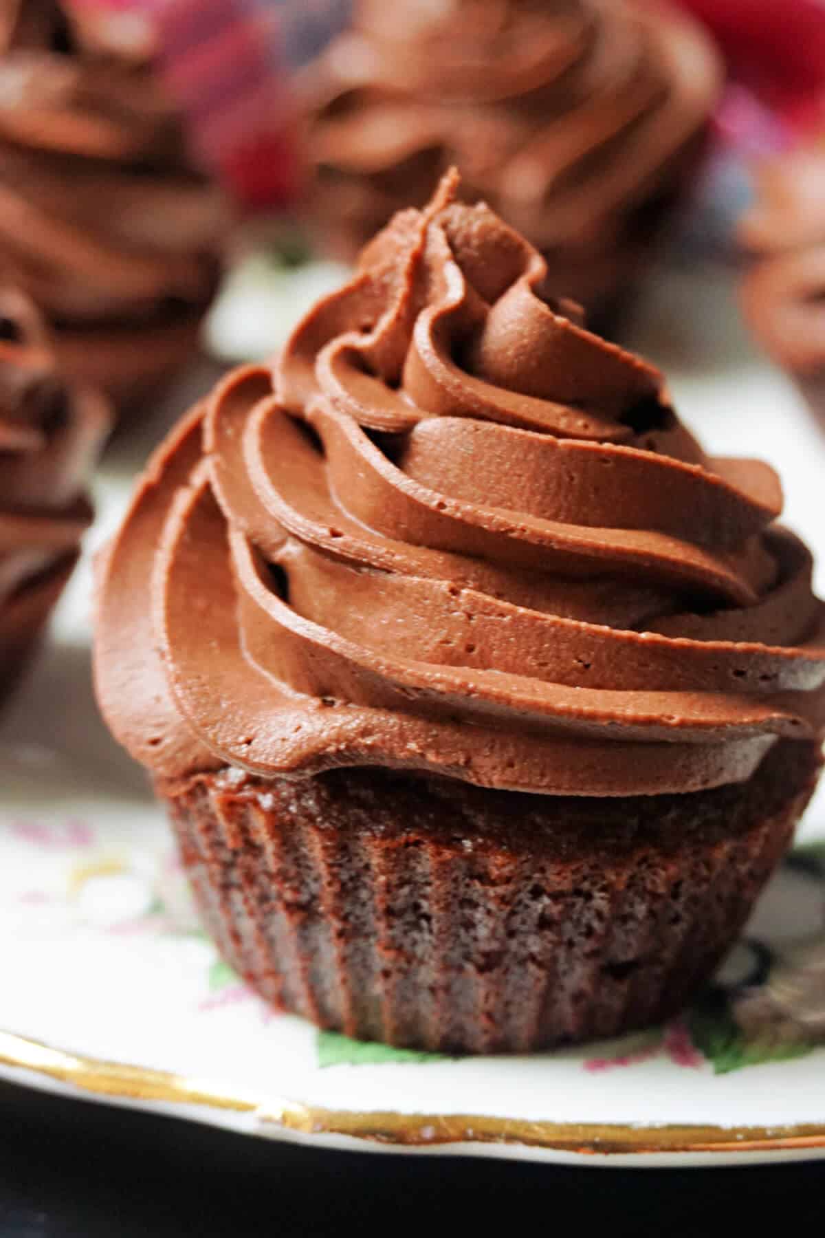 A chocolate cupcake with chocolate buttercream on a white plate with more cupcakes in the background.