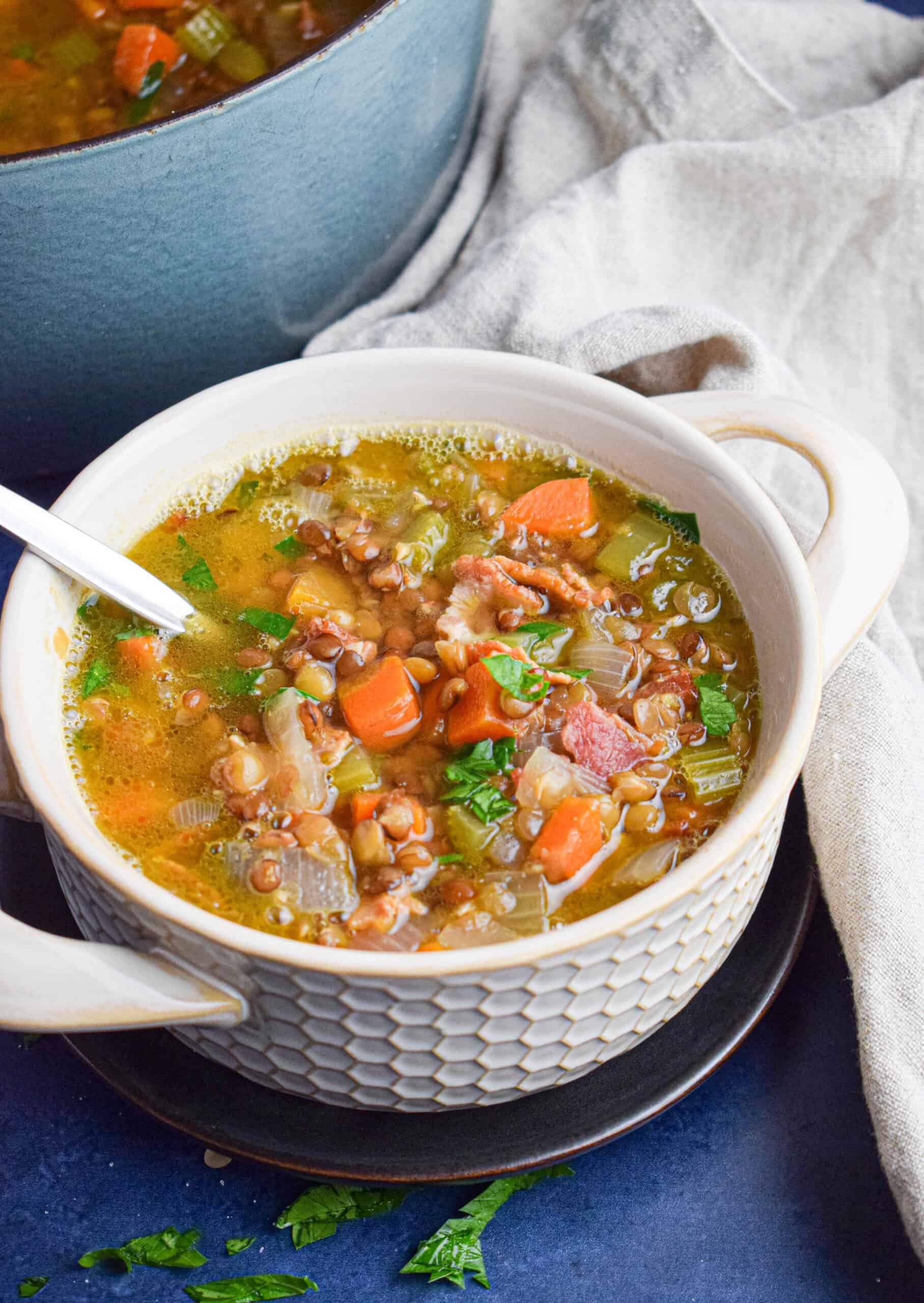 A cream colored bowl is filled with soup with lentils, bacon, carrots, celery and onions topped with parsley next to a tan colored cloth on a blue background.