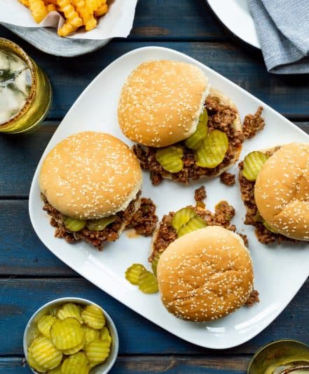 White platter of 4 Heinz Chili Sauce Sloppy Joes with a basket of crinkle cut fries, a bowl of pickle slices, yellow glasses of water, and a stack of plates with denim colored napkins.