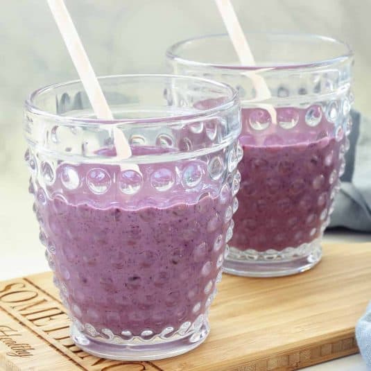 Two glasses of blueberry kefir smoothie with straws. The glasses are sitting on a wood board with a blue napkin in the background.