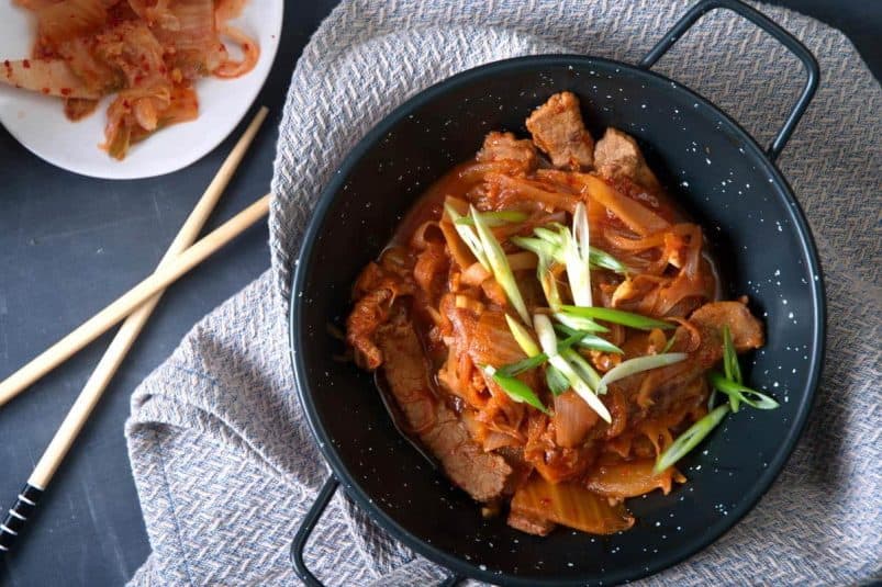 Kimchi stew in a black bowl with sliced scallions. Chopsticks and kimchi on the side.