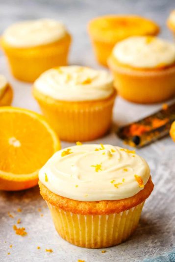 An orange cupcake topped with a swirl of orange cream cheese frosting and garnished with orange zest with oranges and more orange cupcakes in the background.