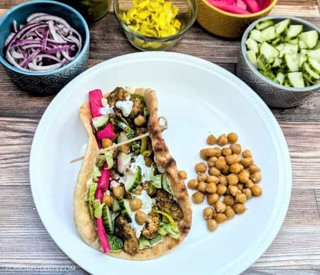 Sheet Pan Za'atar Chicken & Chickpeas in a flatbread with a variety of toppings in the background.
