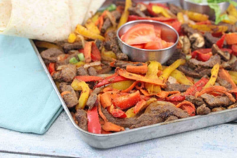 Baking tray with cooked steak, bell peppers, and veggies with a steel bowl of fresh tomatoes in the middle and flour tortillas folded to the side.