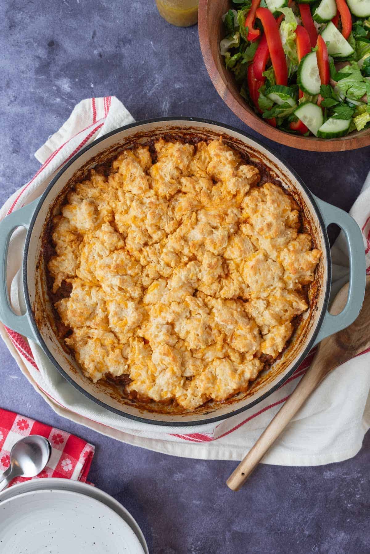 Sloppy Joe Casserole with cheddar biscuit top, ready to serve from a pale green braising pan. A green salad, a wooden serving spoon, plates and napkins beside the dish.