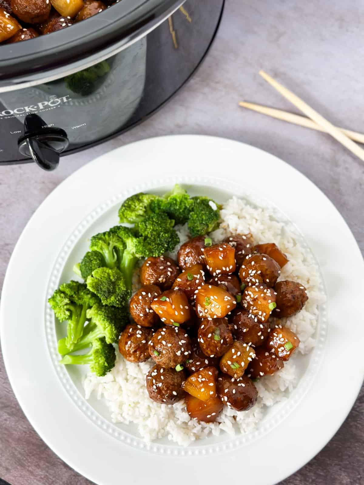 Plate of slow cooker pineapple teriyaki meatballs on rice with broccoli on the side and chopsticks and a slow cooker in the background.