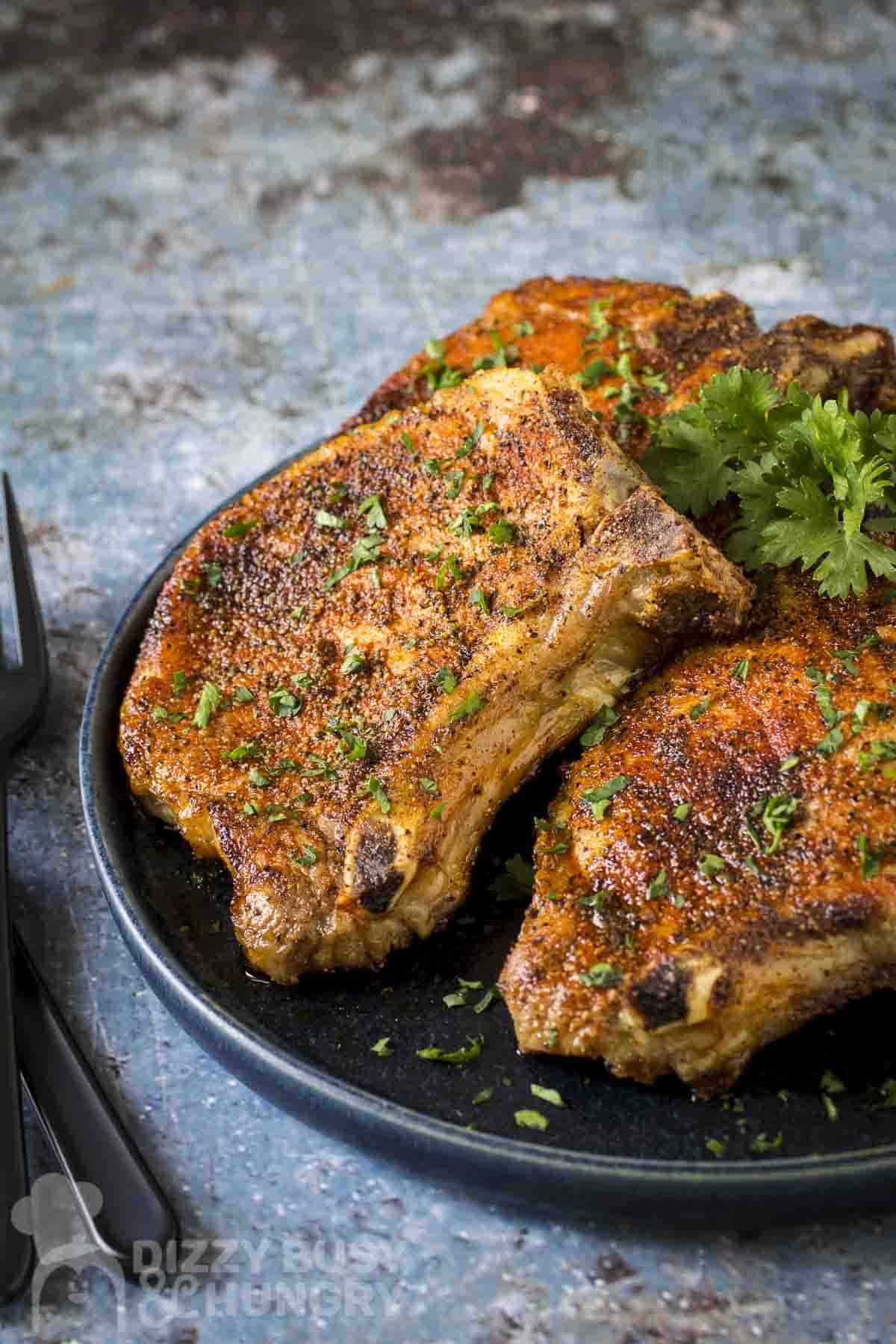 Side shot of three pork chops on a black plate garnished with fresh parsley with a black fork on the side.