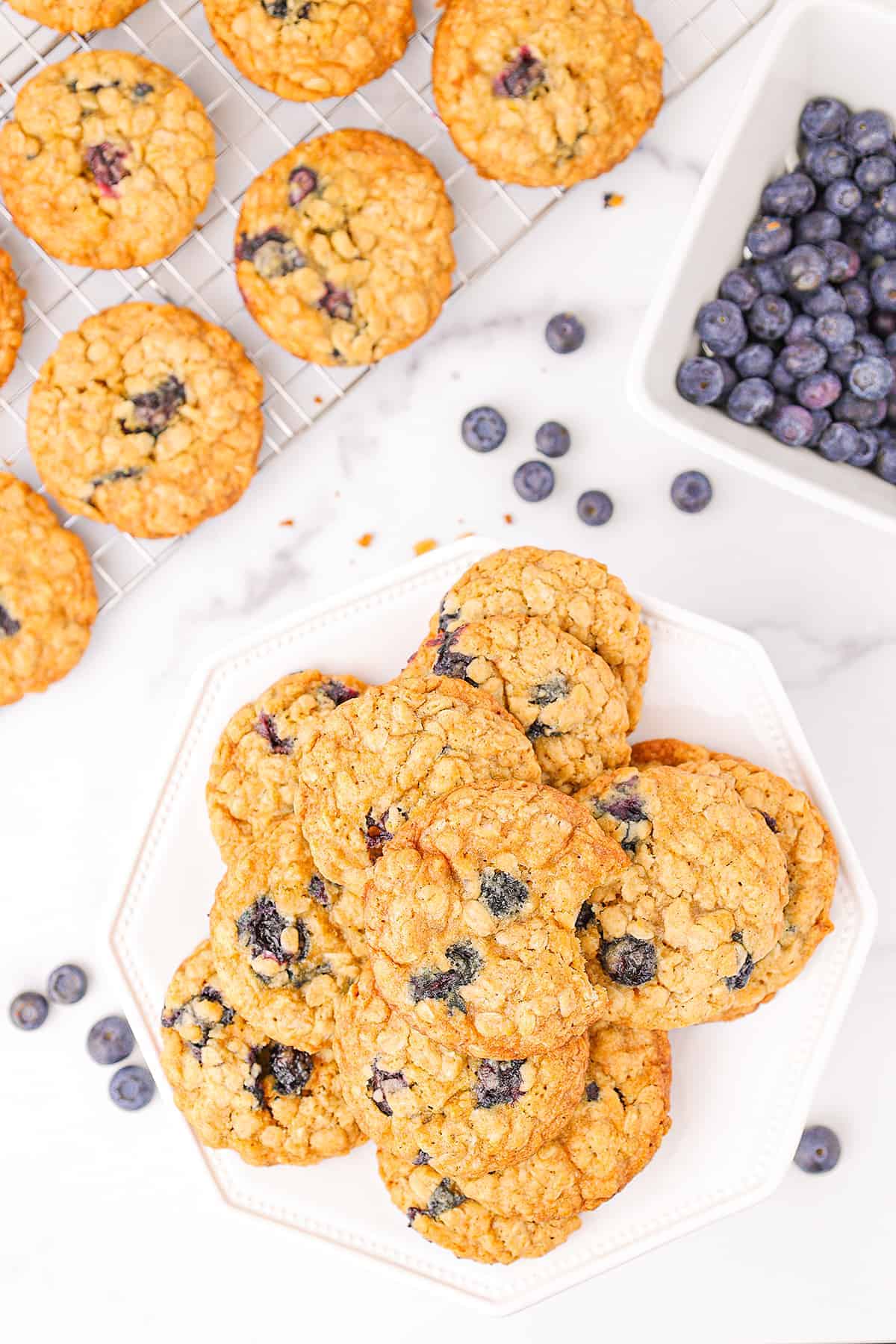 A large stack of lemon blueberry oatmeal cookies on a white cake stand with a white container of fresh blueberries and six more oatmeal cookies on a wire rack.