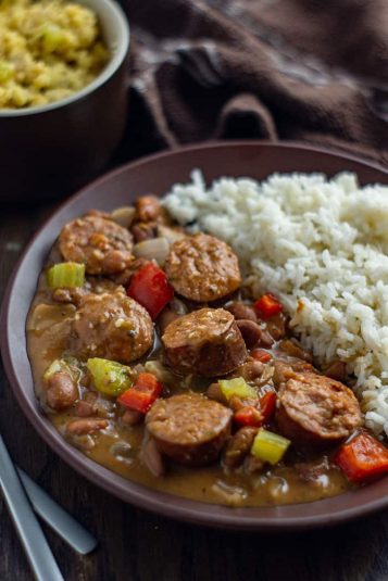 Cooked beans and sausage in plate with rice on the side and cornbread in the background.