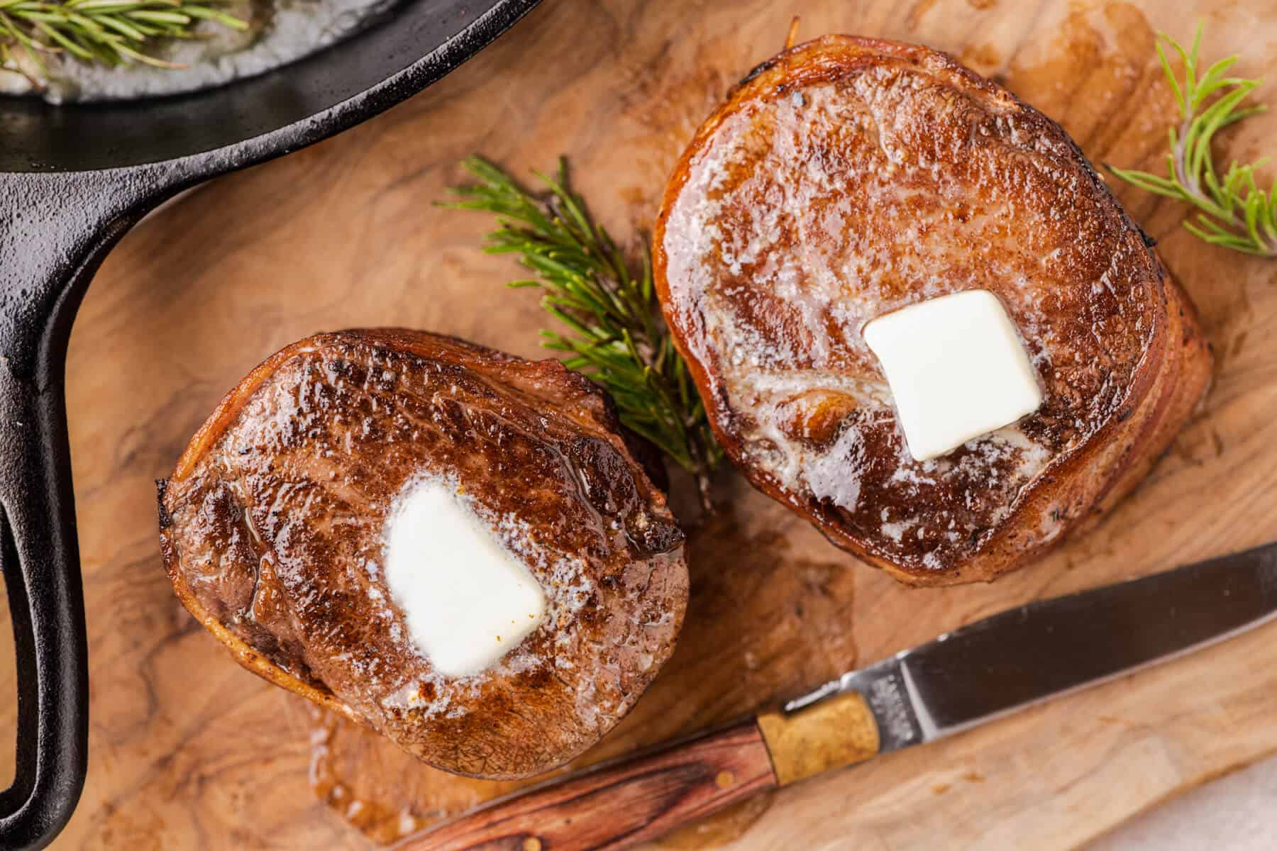 Two perfectly seared bacon wrapped filet mignon steaks garnished with melting butter and fresh rosemary sit on a wooden serving board.