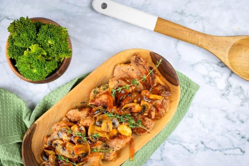 Pork chops with gravy, mushrooms and fresh thyme in serving dish. Wooden spoon and broccoli on the side.