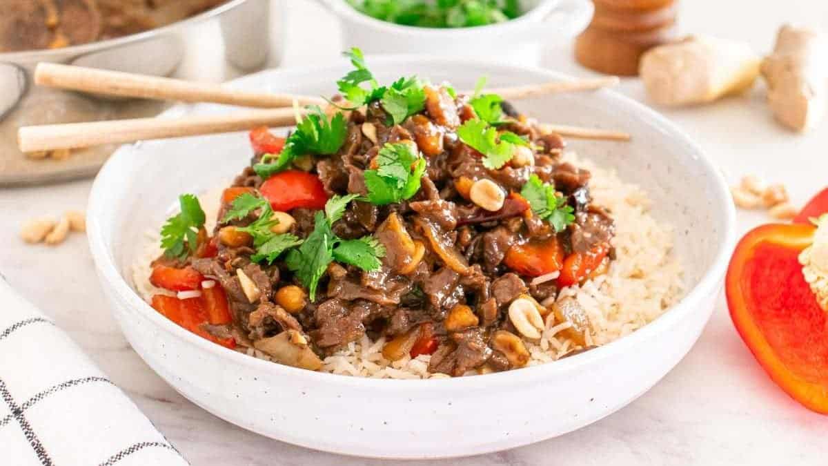 A bowl of rice topped with beef stir-fry, red bell peppers, peanuts, and cilantro. Chopsticks rest on the bowl. Additional ingredients are visible in the background.