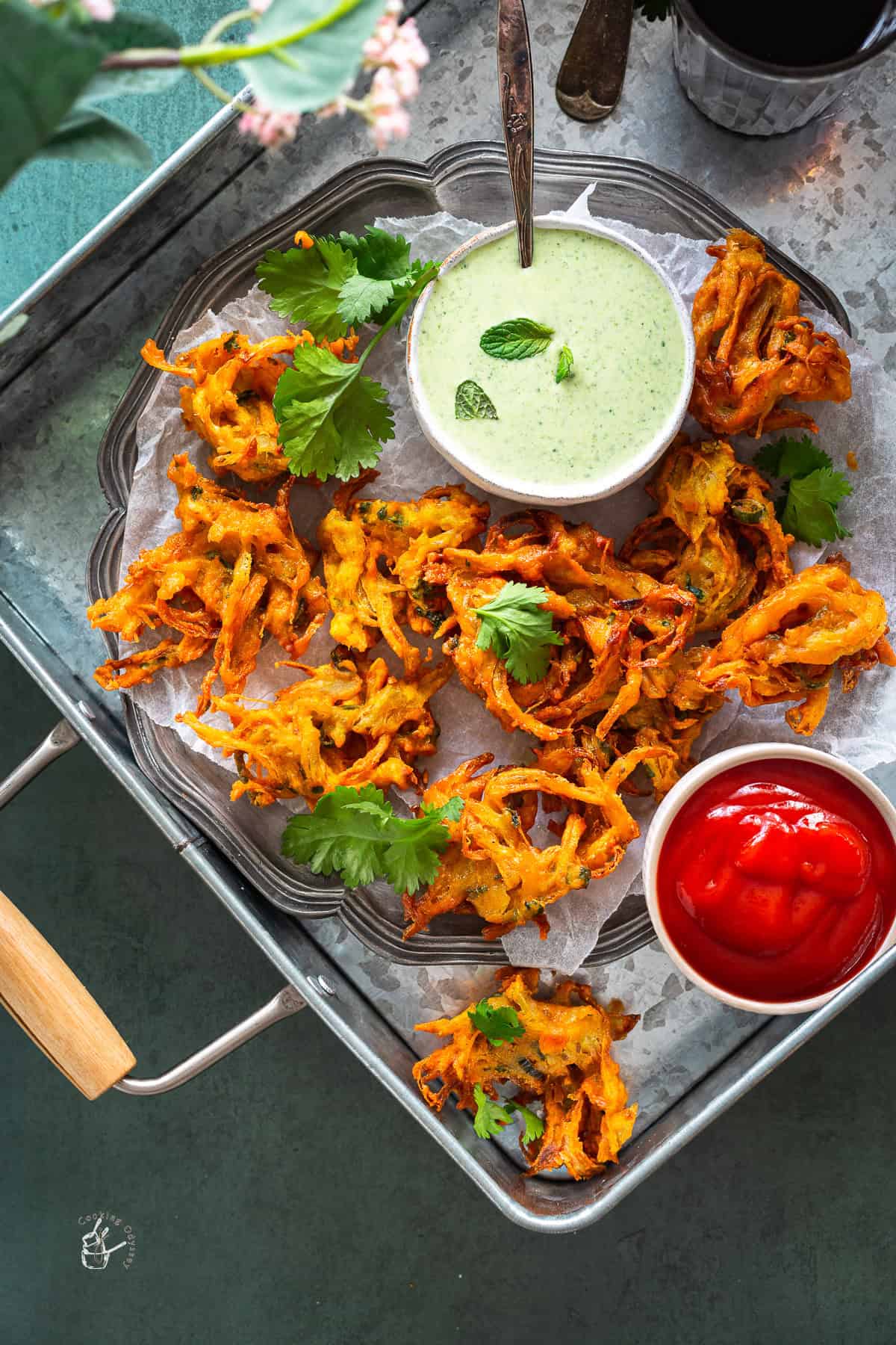 Onion bhaji served on parchment paper on a pewter plate, garnished with fresh coriander leaves next to indian dips bowls.