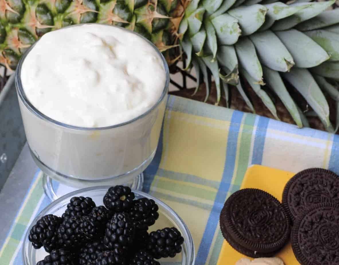 Glass bowl of pineapple dip, blackberries, cookies, and a pineapple in the background.