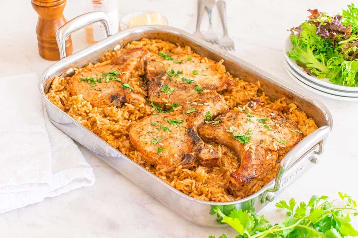 A pork chop and rice casserole sits invitingly in a baking dish, featuring four perfectly cooked chops on a bed of seasoned rice, garnished with parsley. Nearby, a towel, salad bowl, and condiments rest elegantly on the marble surface.