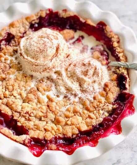 Warm cherry cobbler oozing over the side of a white pie plate with crumble topping and vanilla ice cream melting on top.