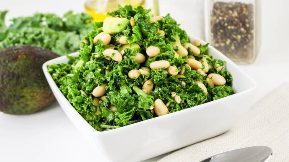 A square white bowl filled with kale salad, featuring beans and avocado pieces, placed on a table with a spoon beside it.
