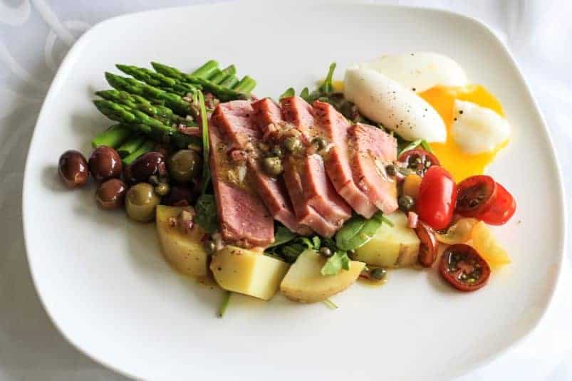 Nicoise salad composed of rare sliced tuna, nicoise olives, and runny egg arranged on a white plate.