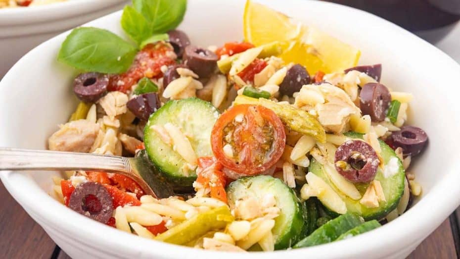 A bowl of Mediterranean-style salad with orzo, cucumber, cherry tomatoes, olives, red peppers, tuna, and a lemon wedge, garnished with basil.