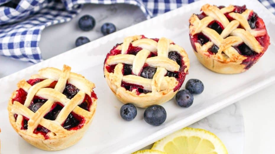 Three mini blueberry pies with lattice tops on a rectangular white plate, surrounded by fresh blueberries and lemon slices, and a blue checkered cloth in the background.