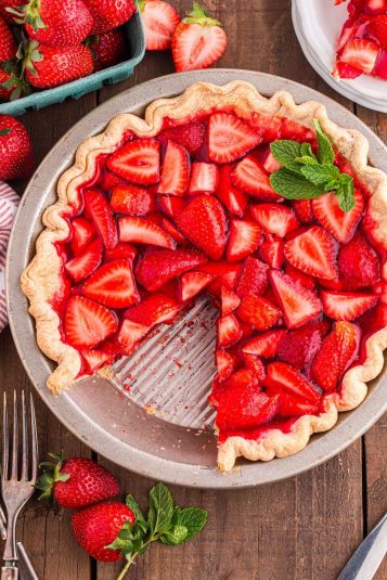 A strawberry pie with a missing slice on a wooden table. Fresh strawberry slices and a mint garnish on top.