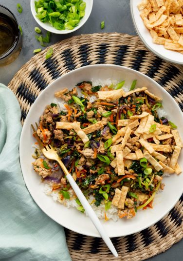 White bowl filled with Egg Roll in a Bowl with Trader Joe's Cruciferous Crunch on top of a black and natural wicker placement, a light blue napkin, bowl of sliced green onions, a bowl of wonton strips, and a gray glass of water.