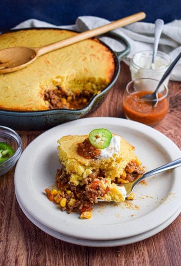 Cast iron skillet of cooked tamale casserole and in front is a plated serving of the tamale pie with sour cream, salsa and a sliced jalapeno.