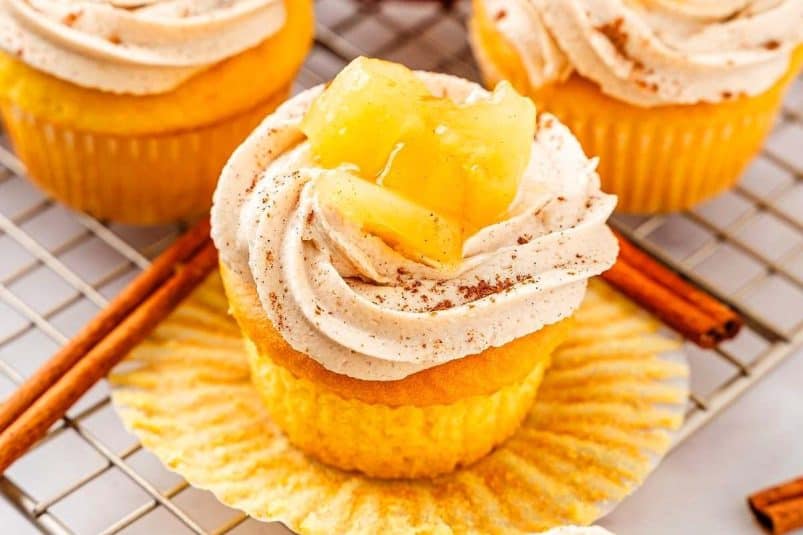 An apple pie cupcake is topped with whipped cream and apple slices on a cooling rack. A cinnamon stick lies in front, while blurred red apples set the background tone.