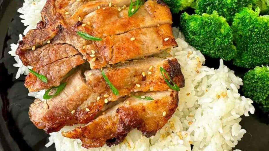 Grilled pork slices on white rice, garnished with sesame seeds and green herbs, served with broccoli florets.