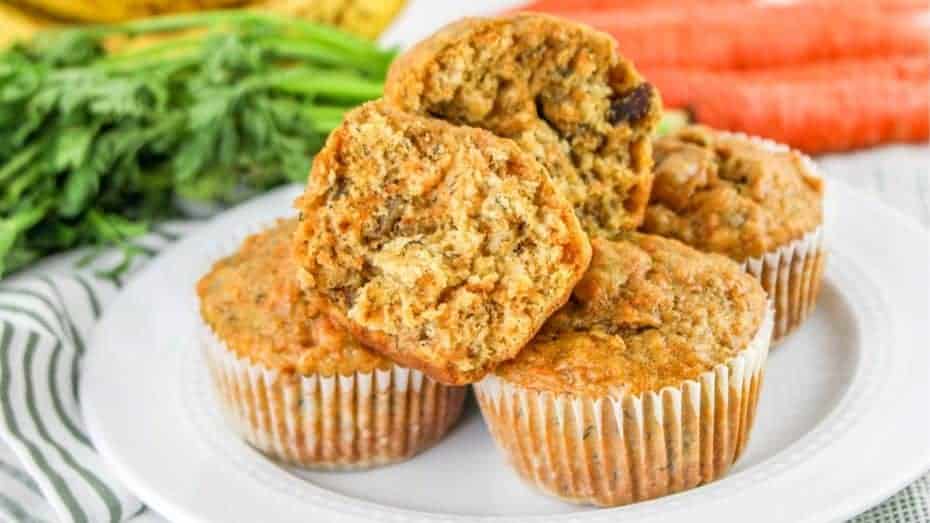 A stack of carrot banana muffins on a white plate, surrounded by fresh carrots and a striped cloth.
