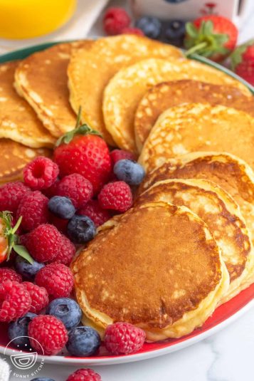 A large red round platter holding pan fried cottage cheese pancakes that are arranged in a circle around the plate. In the center are fresh mixed berries.