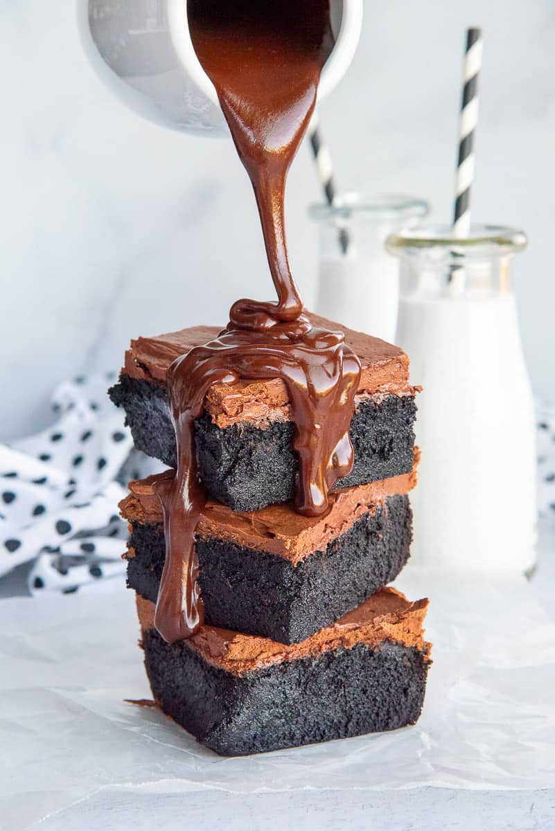 Chocolate ganache pours down the side of a stack of dark chocolate fudgy brownies.