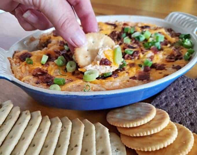 Image shows a hand dipping a cracker into Hot Bacon Cheddar Dip in a ceramic dish.